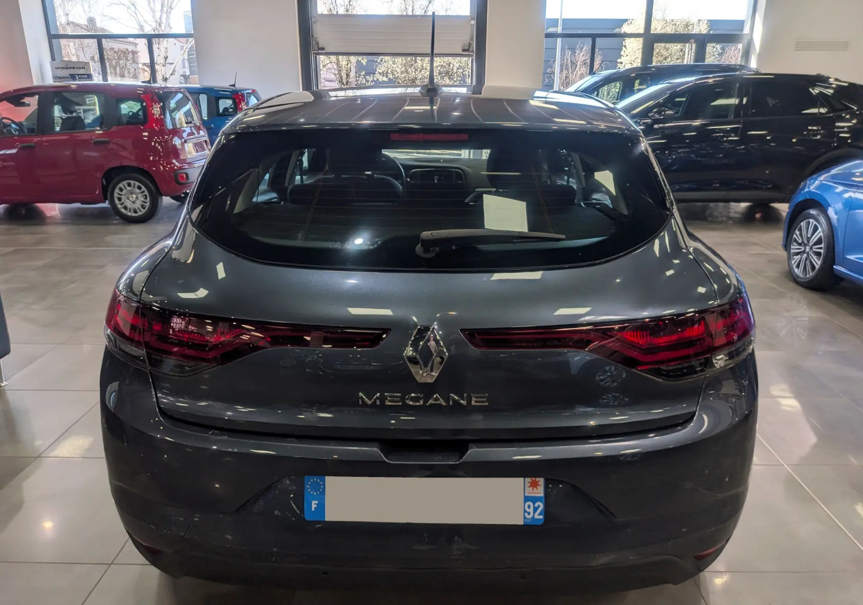 Vue arrière d'une Renault Mégane IV Berline gris titanium 2022, avec feux arrière LED et logo central visible en showroom.