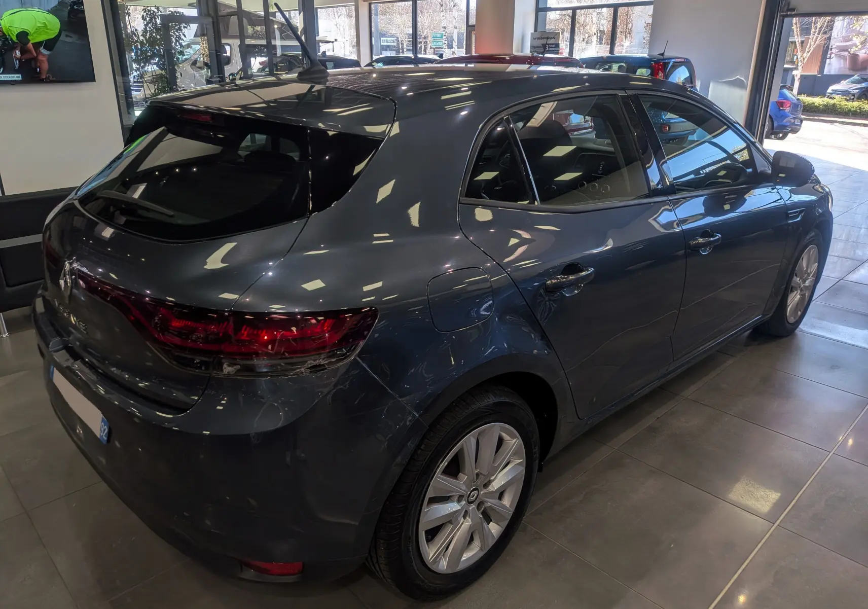 Vue 3/4 arrière droit d'une Renault Mégane IV Berline gris Titanium en showroom, mettant en valeur ses feux arrière et jantes.