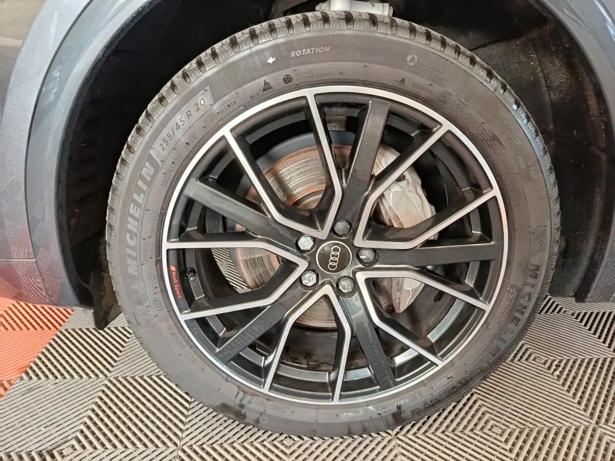 Gros plan sur la roue avant droite d’un Audi Q5 gris clair 2023, jante noire à détails argentés avec logo Audi au centre.