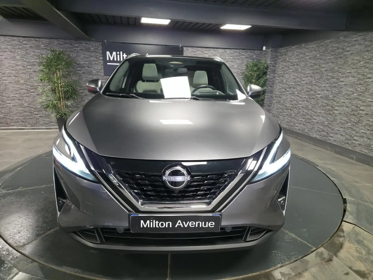 Vue avant d'un Nissan Qashqai 1.5 e-Power gris métallisé avec phares LED allumés en intérieur showroom.