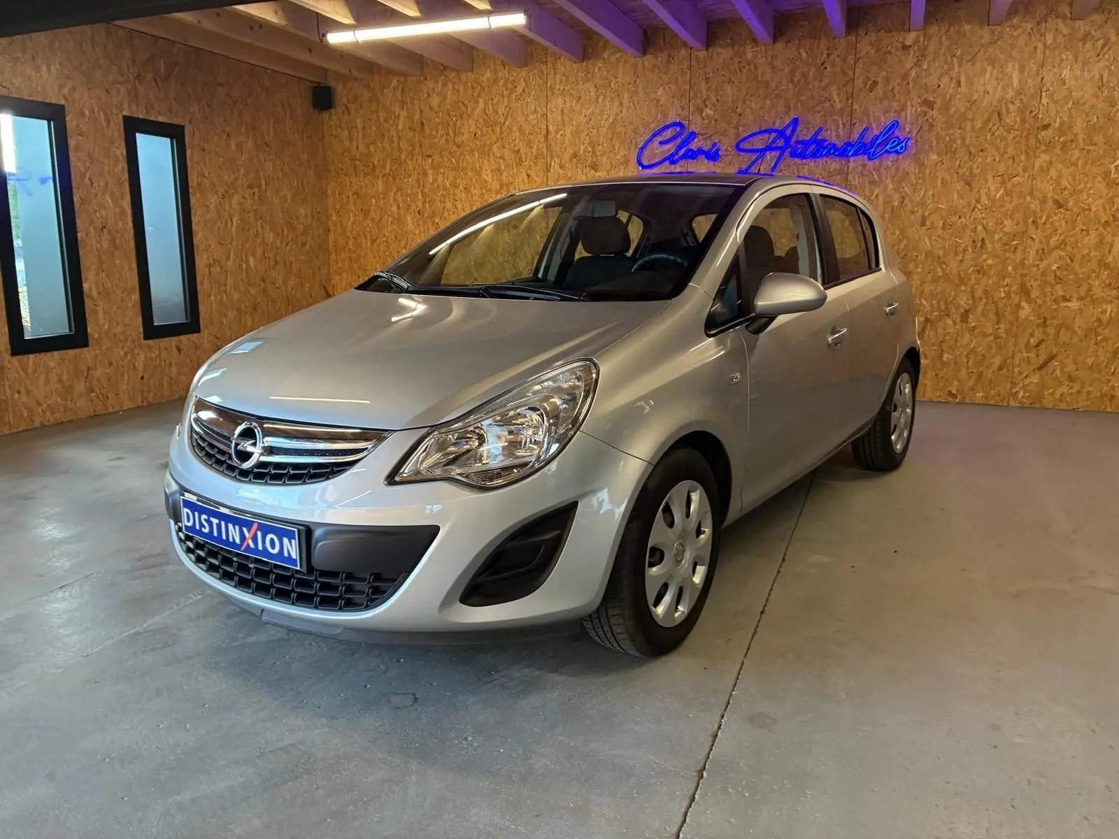 Opel Corsa gris clair vue 3/4 avant droit dans un showroom avec mur en bois et enseigne lumineuse bleue.