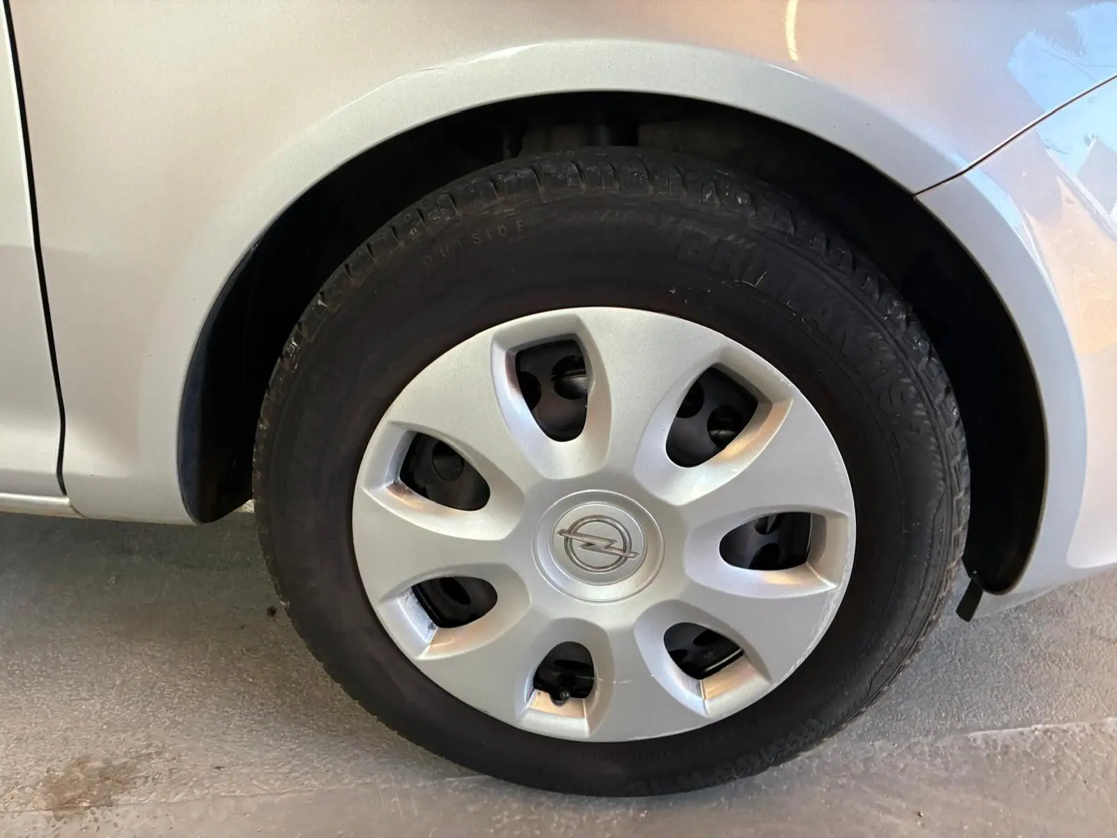 Gros plan sur la roue avant gauche d’une Opel Corsa gris clair, avec enjoliveur argenté et logo Opel au centre.