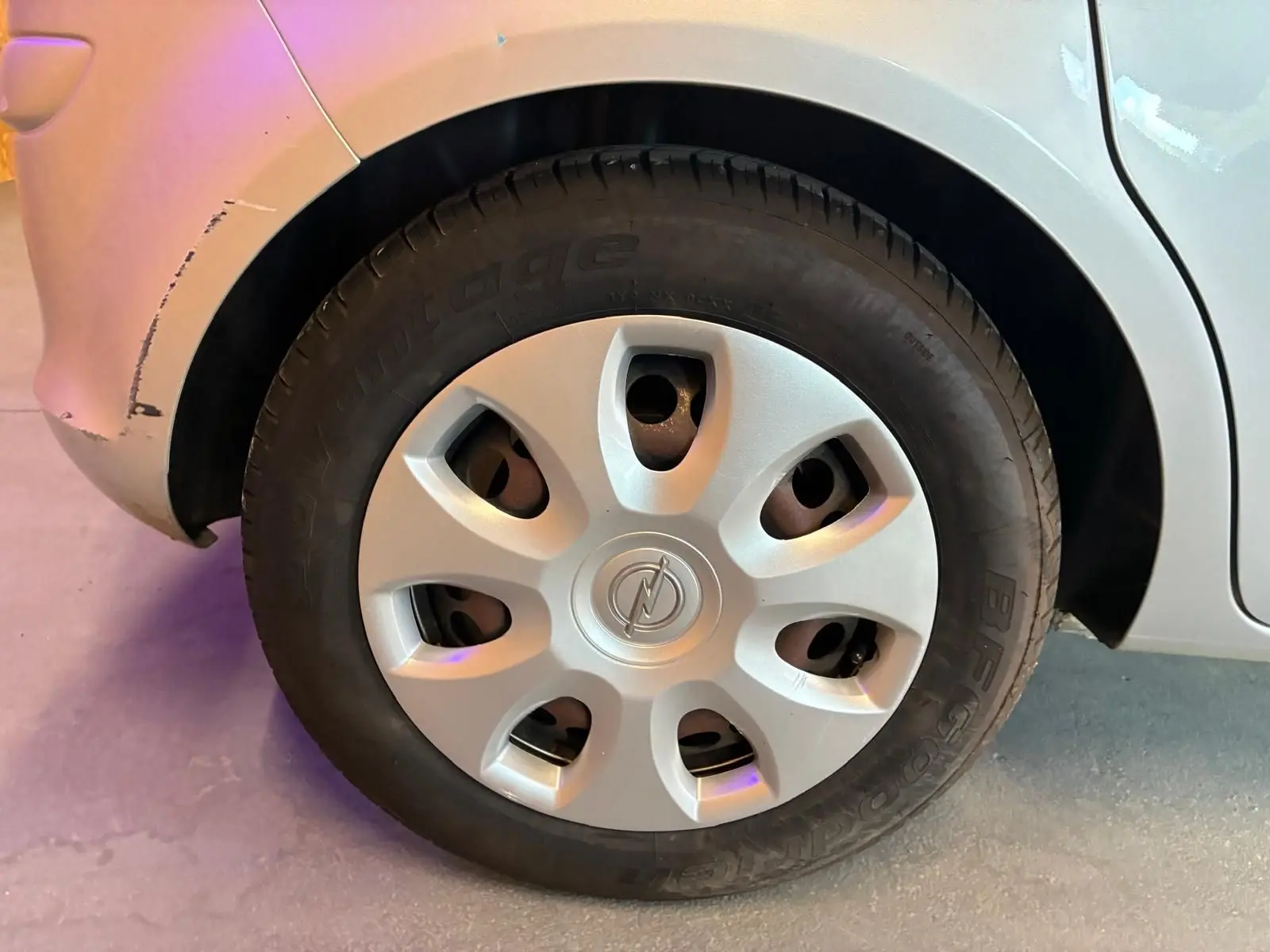 Gros plan sur la roue arrière droite d’une Opel Corsa gris clair de 2012 avec enjoliveur d’origine et carrosserie légèrement éraflée.