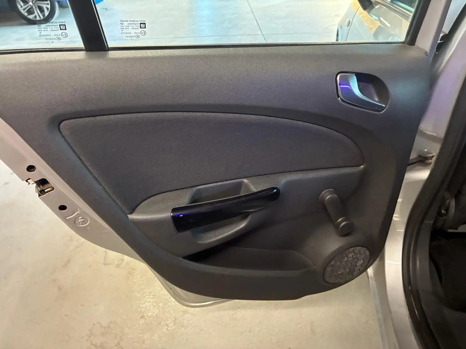 Vue intérieure de la porte arrière gauche grise claire de l'Opel Corsa 2012 avec poignée noire et manivelle de vitre.