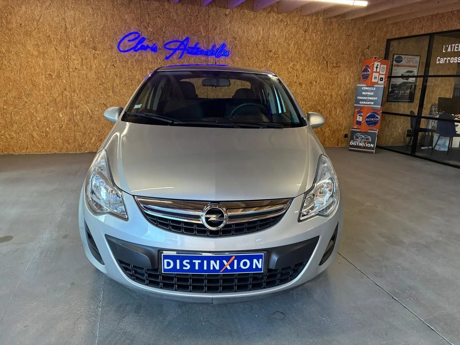Vue de face d'une Opel Corsa gris clair 2012, avec calandre chromée et plaque Distinxion, en intérieur showroom.
