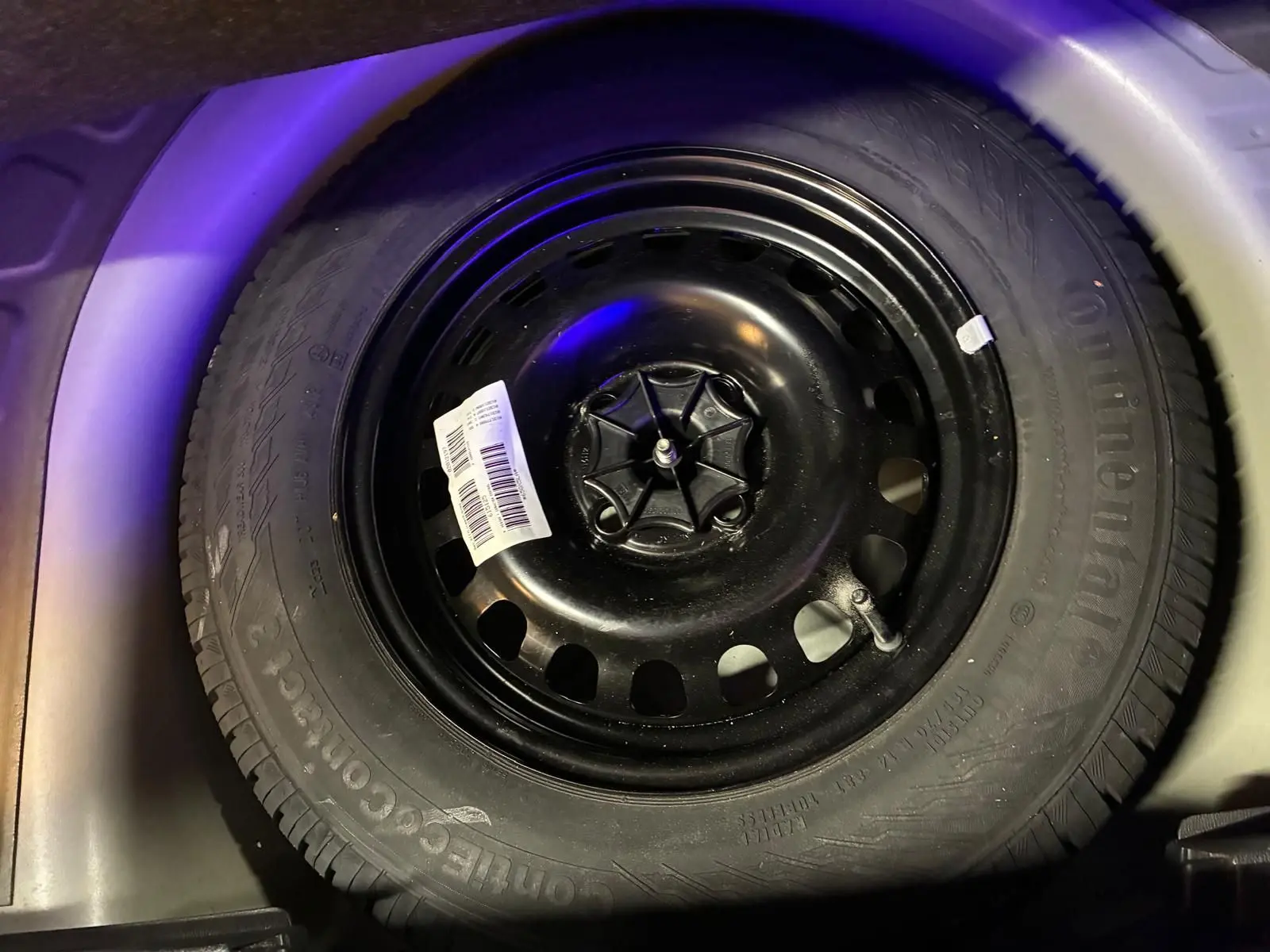 Gros plan sur la roue de secours noire d'une Opel Corsa gris clair, visible dans le coffre sous un éclairage tamisé.