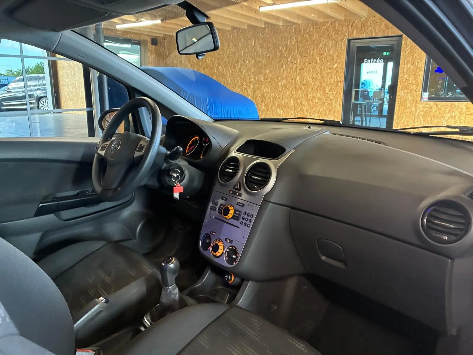 Vue intérieure côté conducteur de l’Opel Corsa gris clair 2012, tableau de bord et volant avec levier de vitesse manuel visibles.