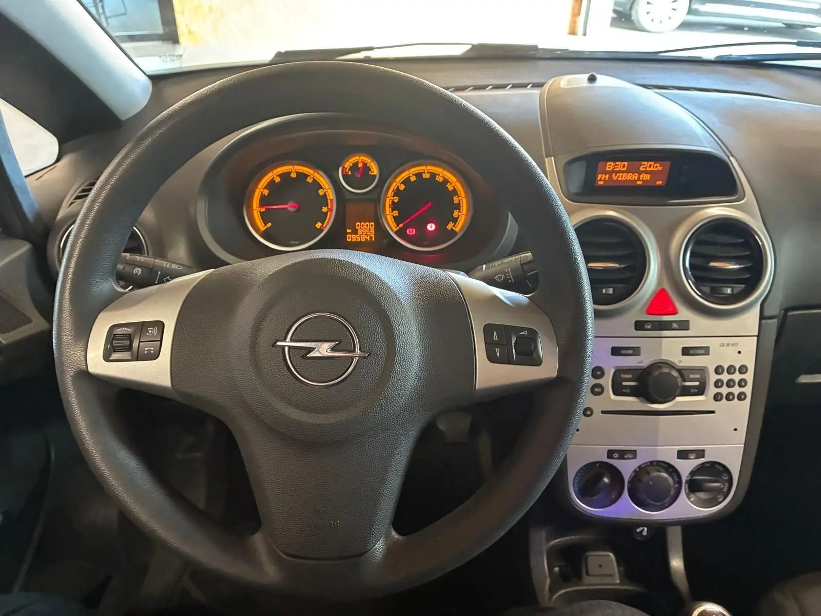 Vue intérieure centrée sur le volant noir d’une Opel Corsa 2012 avec tableau de bord et console centrale gris clair.