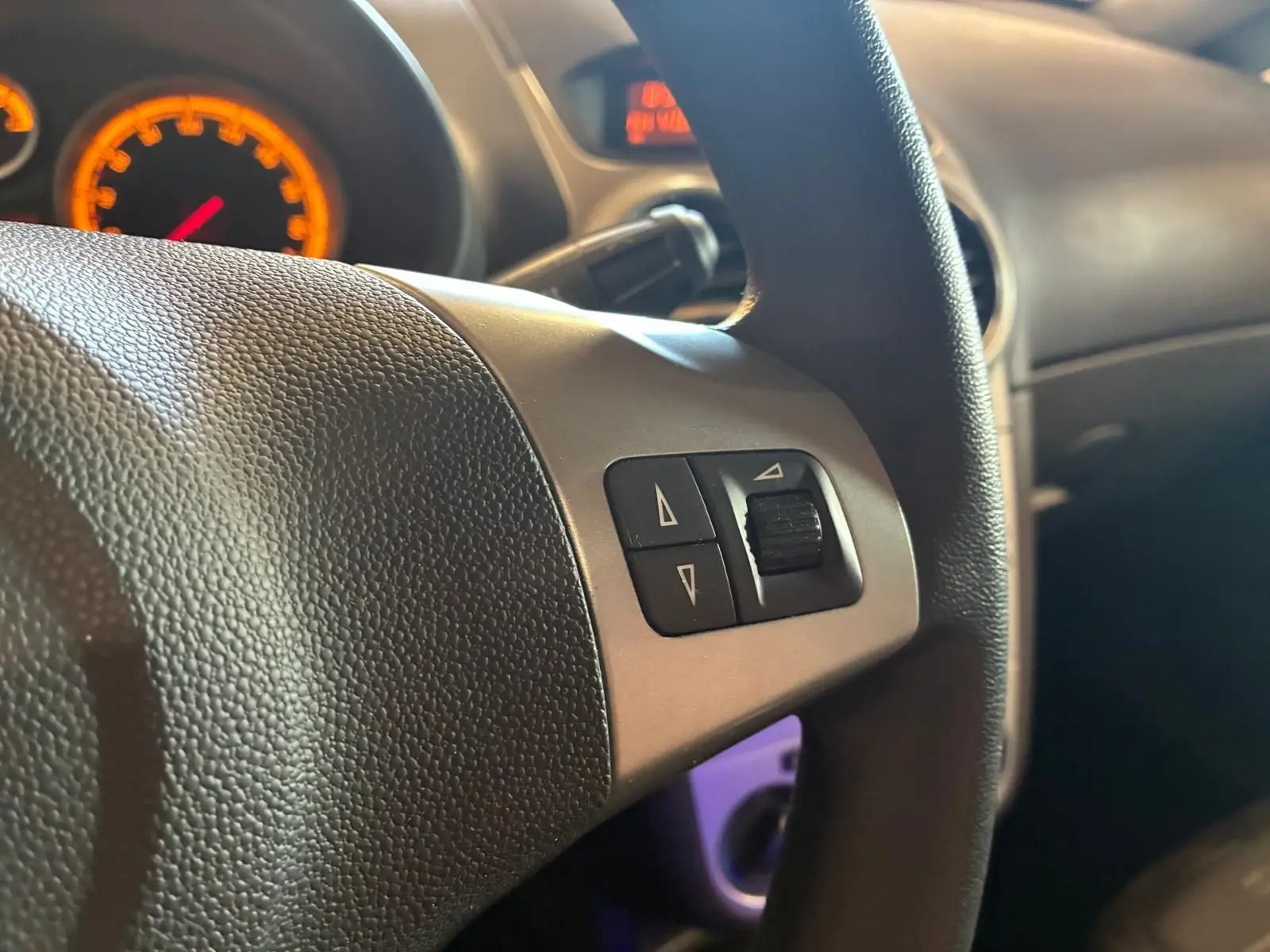 Gros plan sur le volant noir de l’Opel Corsa gris clair 2012, avec commandes intégrées et tableau de bord éclairé en orange.