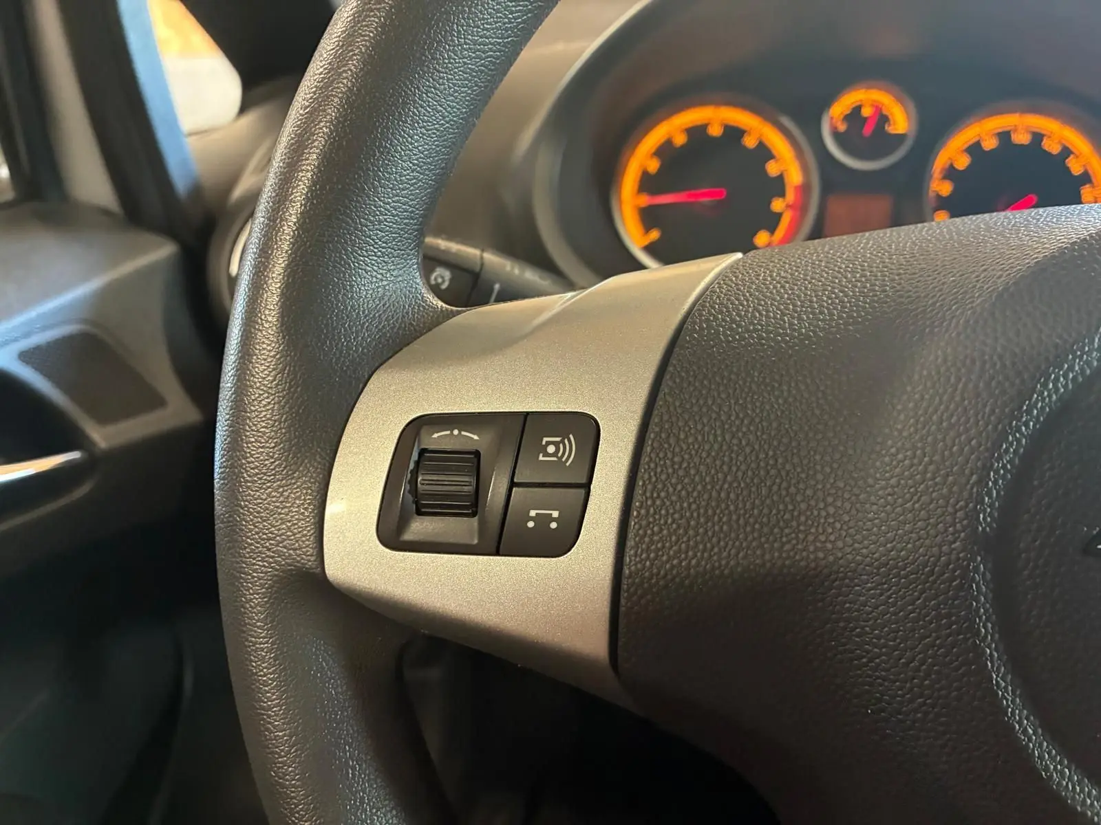 Gros plan sur le volant noir de l'Opel Corsa 2012, avec commandes intégrées et tableau de bord éclairé en orange.