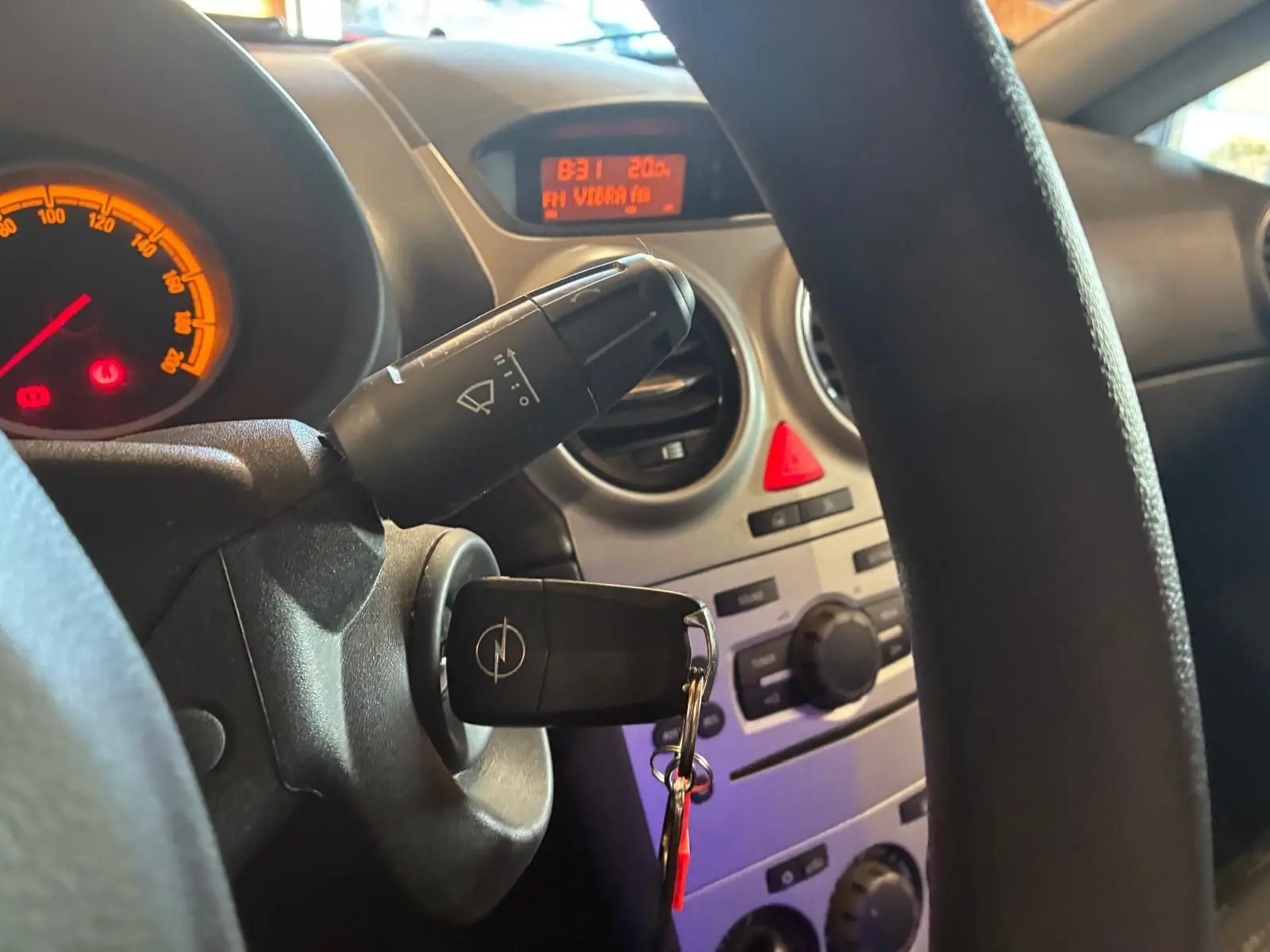 Vue intérieure centrée sur le volant et le tableau de bord d'une Opel Corsa 2012 avec clé insérée et affichage digital orange.