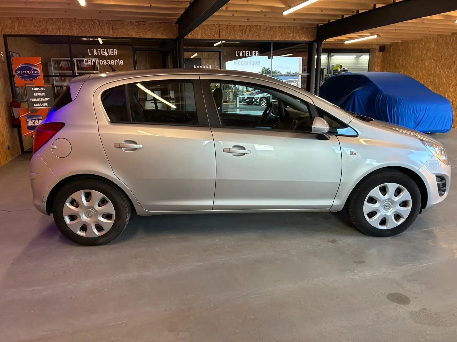 Vue de profil côté gauche d'une Opel Corsa gris clair 5 portes de 2012 stationnée en intérieur, avec jantes acier et enjoliveurs.