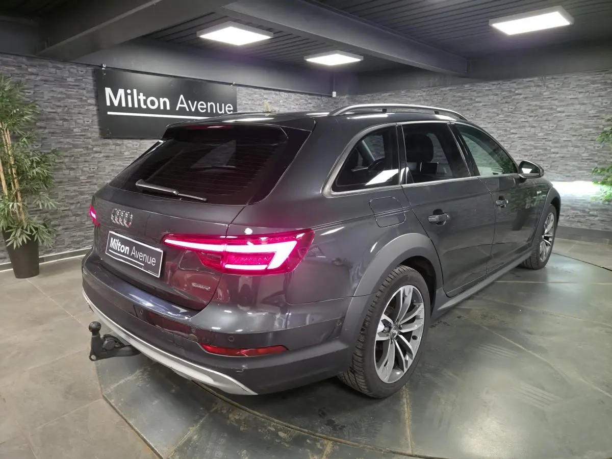 Audi A4 Allroad gris vue 3/4 arrière droit avec feux allumés et crochet d'attelage visible en intérieur showroom.
