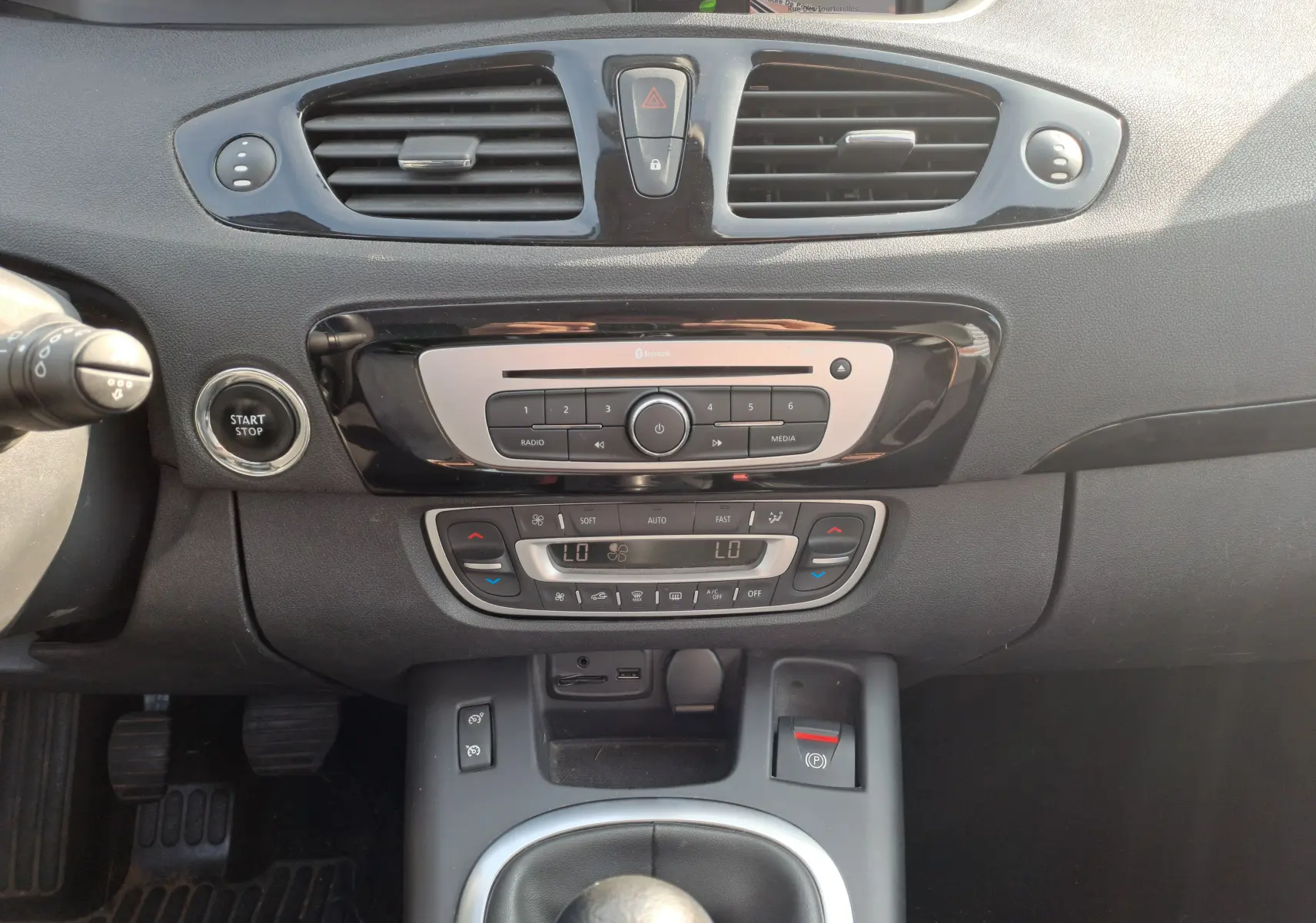 Vue rapprochée du tableau de bord noir du Renault Scenic III 2016, montrant la console centrale avec commandes climatisation et bouton start-stop.