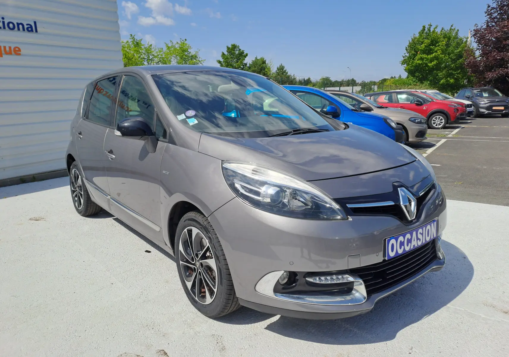 Renault Scenic III gris en 3/4 avant droit, avec jantes alliage et feux avant LED visibles sur parking extérieur.