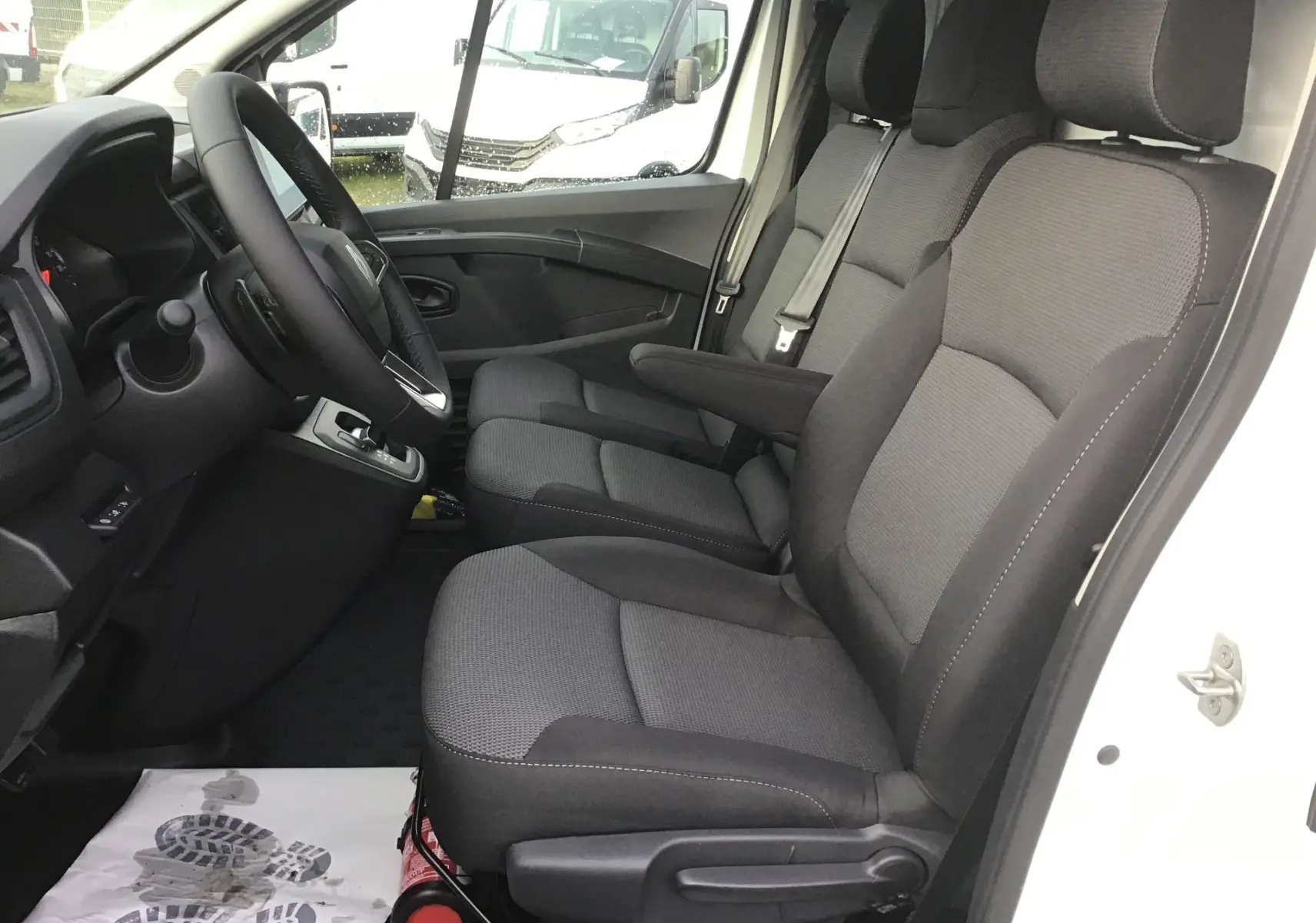 Vue intérieure côté conducteur du Renault Trafic Fourgon blanc 2025, montrant les sièges gris et le volant avec boîte automatique.