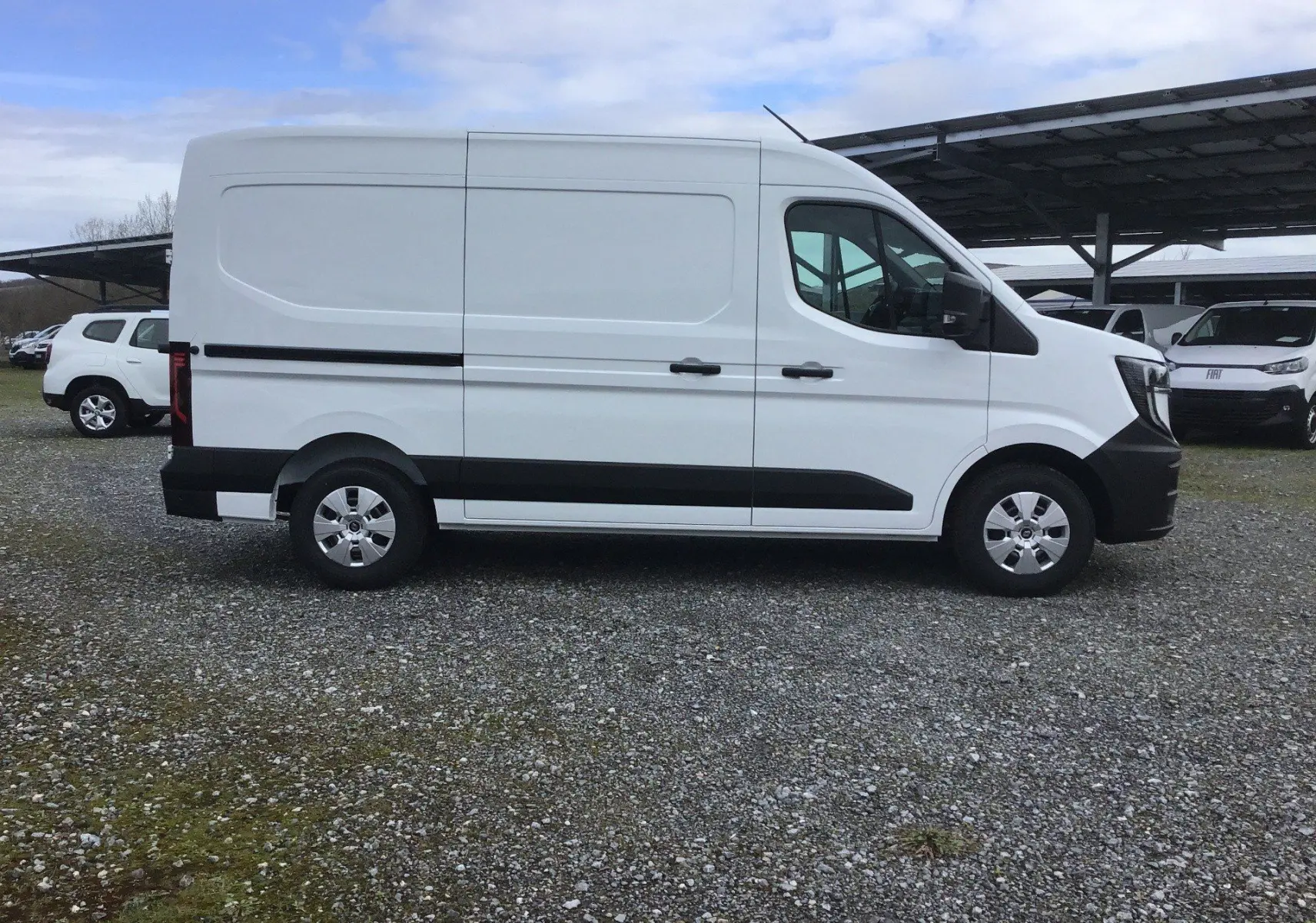 Vue de profil côté droit d’un utilitaire Renault Master blanc avec bandes noires et jantes acier, stationné sur un sol gravillonné.