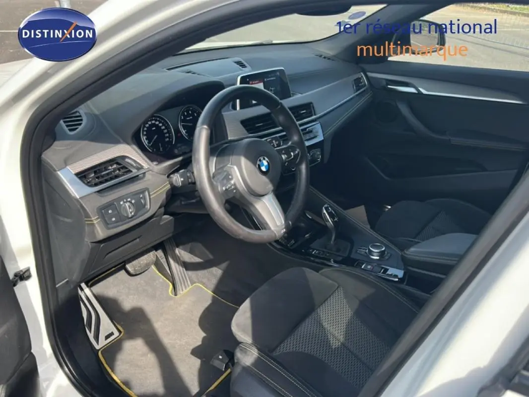Intérieur côté conducteur de BMW X2 blanc, volant sport, console centrale avec boîte auto et écran GPS visible.
