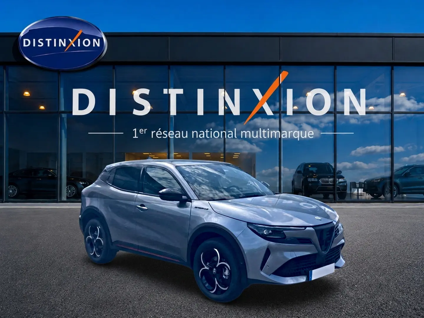 Vue 3/4 avant droite d'une Alfa Romeo Junior gris métallisé devant une vitrine Distinxion, avec jantes en alliage noires et détails noirs.