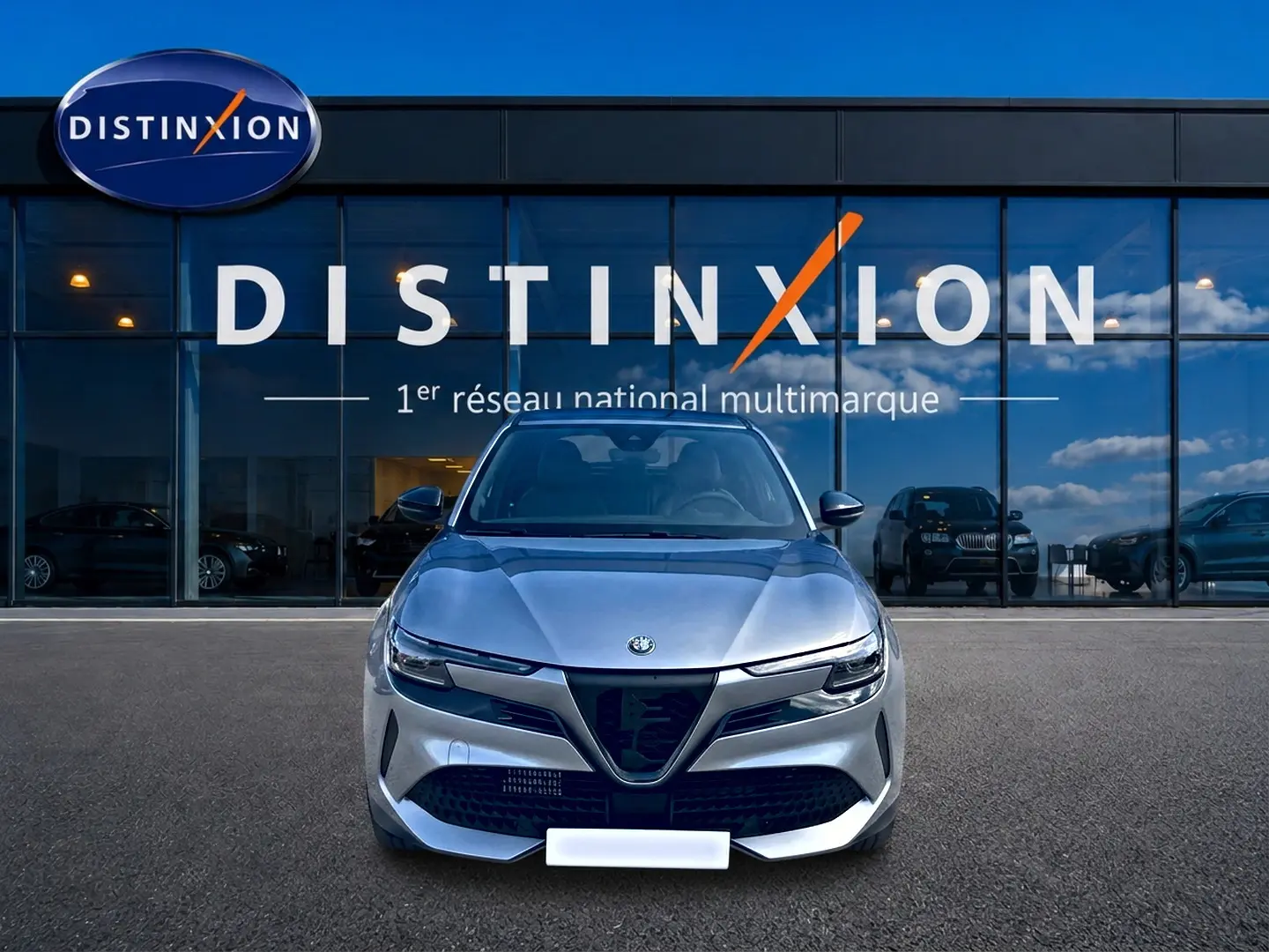 Vue frontale d'une Alfa Romeo Junior 1.2 hybride gris métallisé devant un showroom Distinxion avec reflets de ciel bleu.