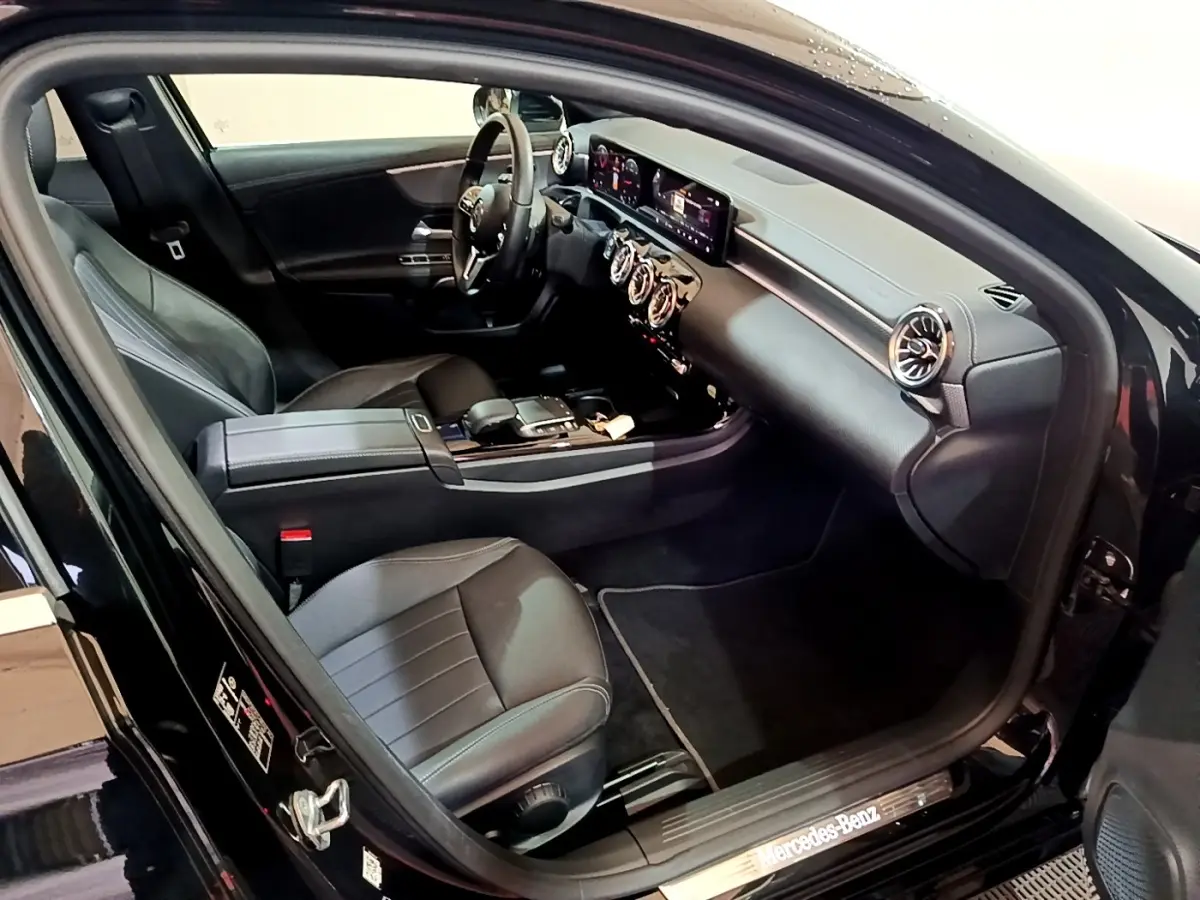 Vue intérieure côté conducteur d'une Mercedes Classe A berline noire, avec tableau de bord digital et aérateurs ronds chromés.