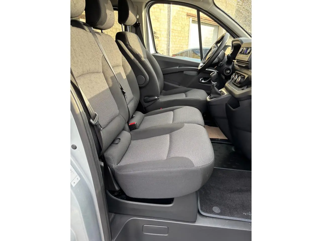 Vue latérale intérieure côté conducteur du Renault Trafic Fourgon gris clair, montrant les sièges avant et le tableau de bord.