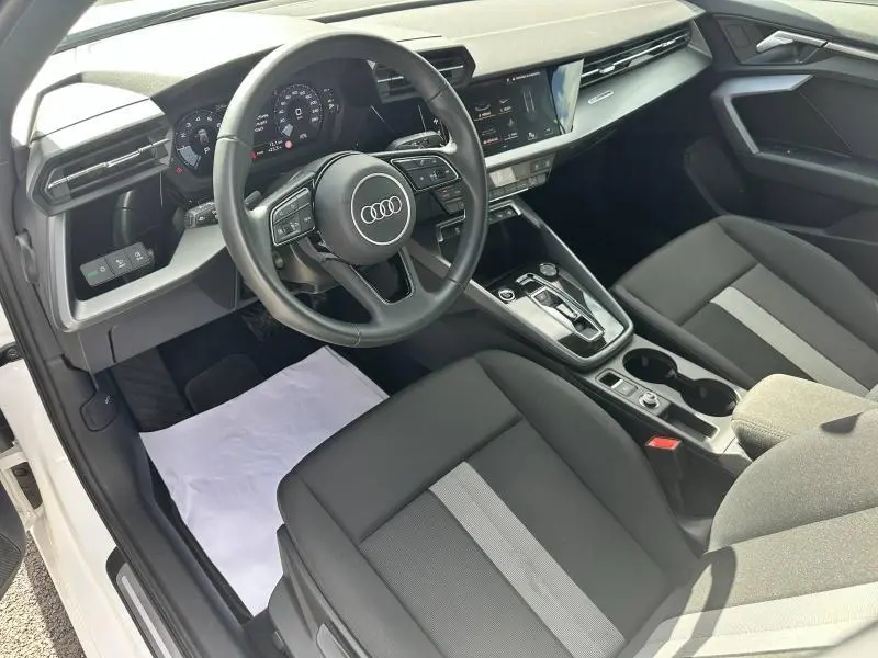 Vue intérieure avant droite de l'Audi A3 Sportback blanc Arkona, montrant le volant cuir multifonction et la console centrale moderne.