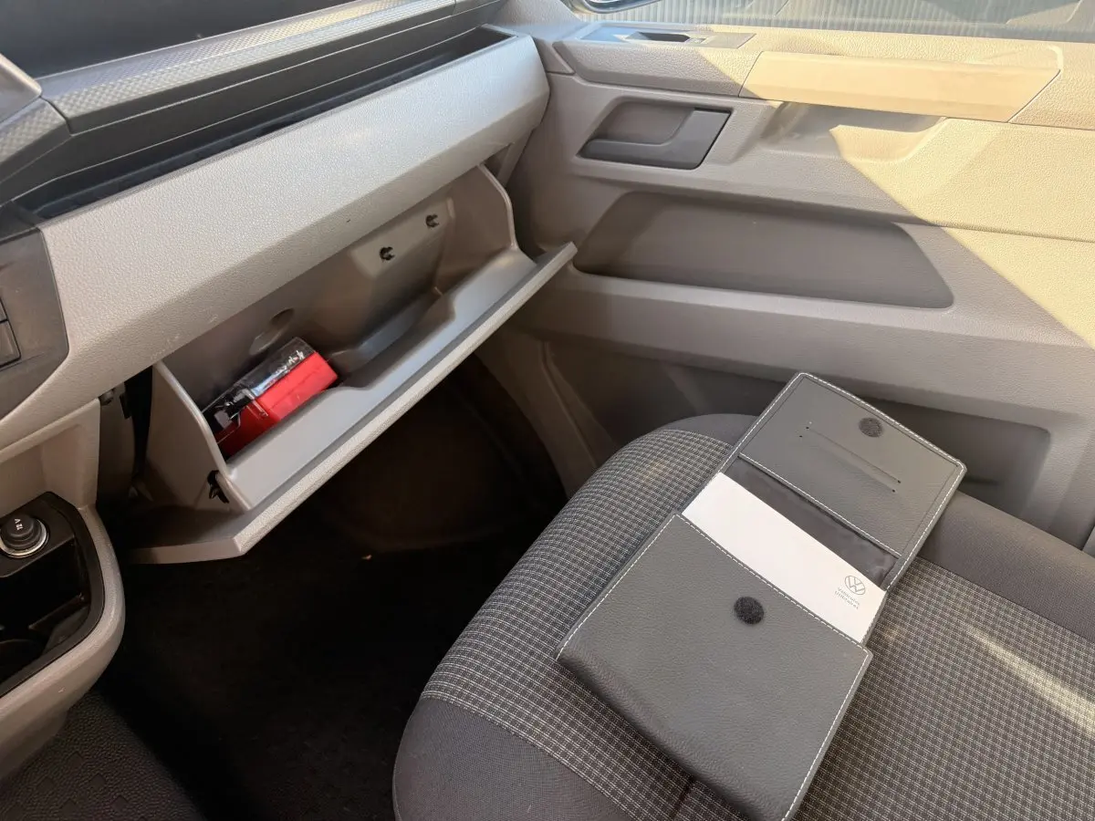 Vue intérieure côté passager du Volkswagen Transporter gris clair 2022, boîte à gants ouverte et pochette sur siège tissu gris.