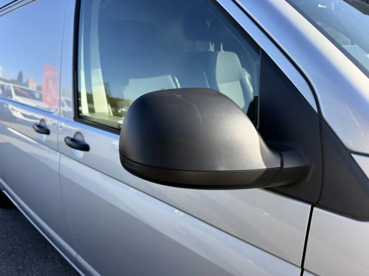 Vue rapprochée du rétroviseur côté conducteur d’un Volkswagen Transporter gris clair 2022, avec portières visibles.