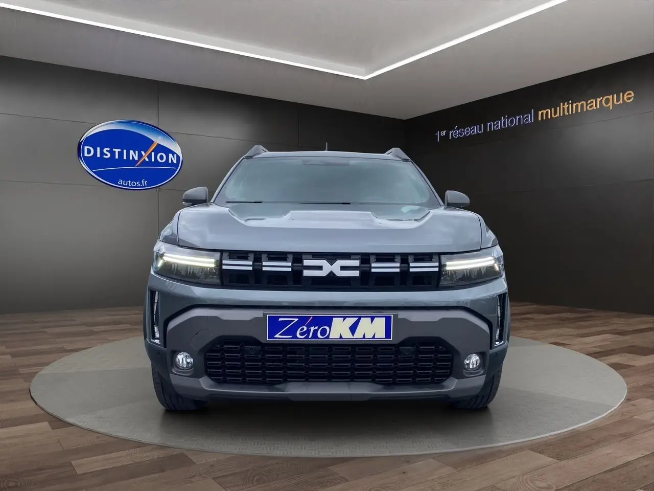 Vue frontale du Dacia Duster 2025 gris schiste, avec calandre noire et phares LED allumés en showroom.