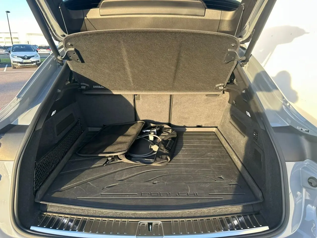 Coffre ouvert vu de l'arrière d'un Porsche Cayenne Coupé 2024 gris arctique avec tapis de coffre et câble de recharge hybride.