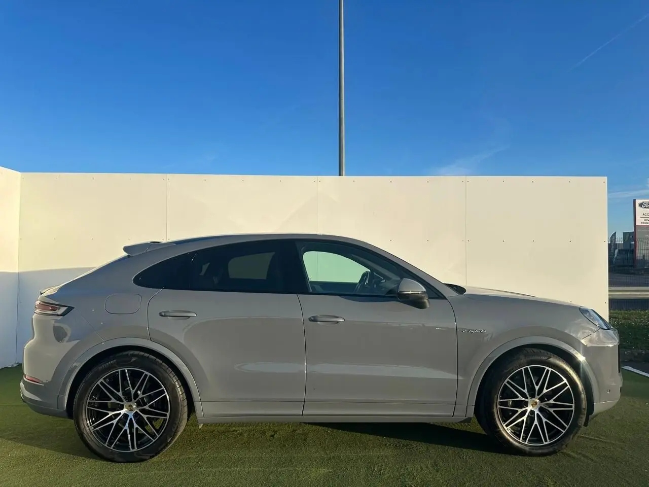 Vue de profil côté gauche d'un Porsche Cayenne Coupé 2024 gris arctique avec jantes noires et badge hybride visible.