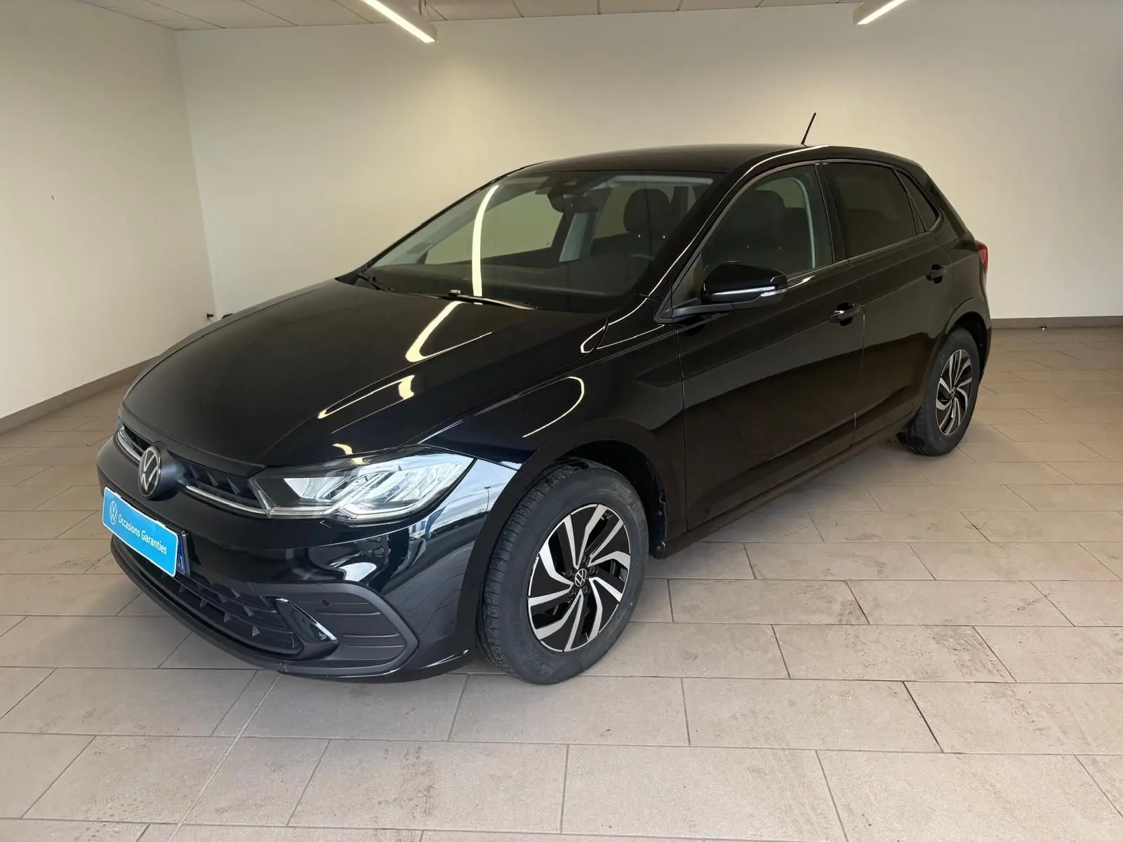 Volkswagen Polo noir intense vue 3/4 avant droit, jantes alliage et calandre chromée visible en intérieur showroom.
