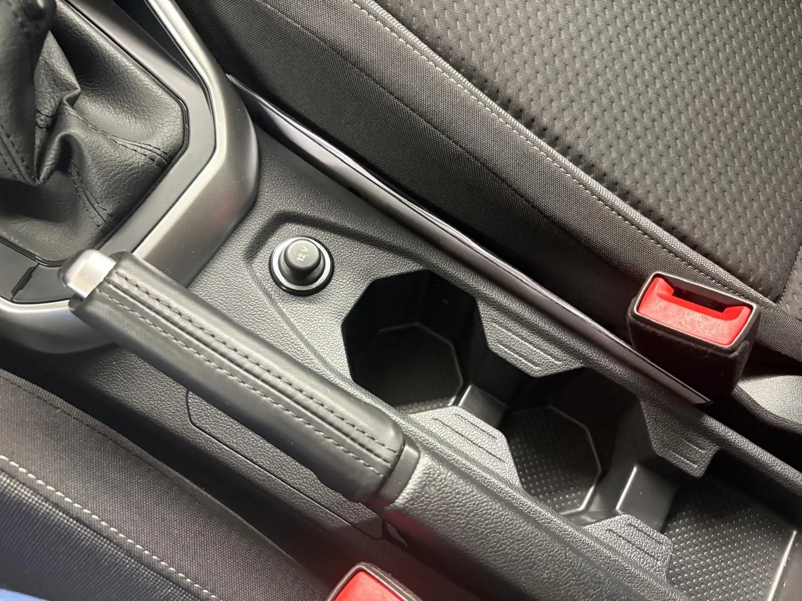 Gros plan intérieur sur le frein à main en cuir, prise 12V et porte-gobelets entre les sièges avant noirs.