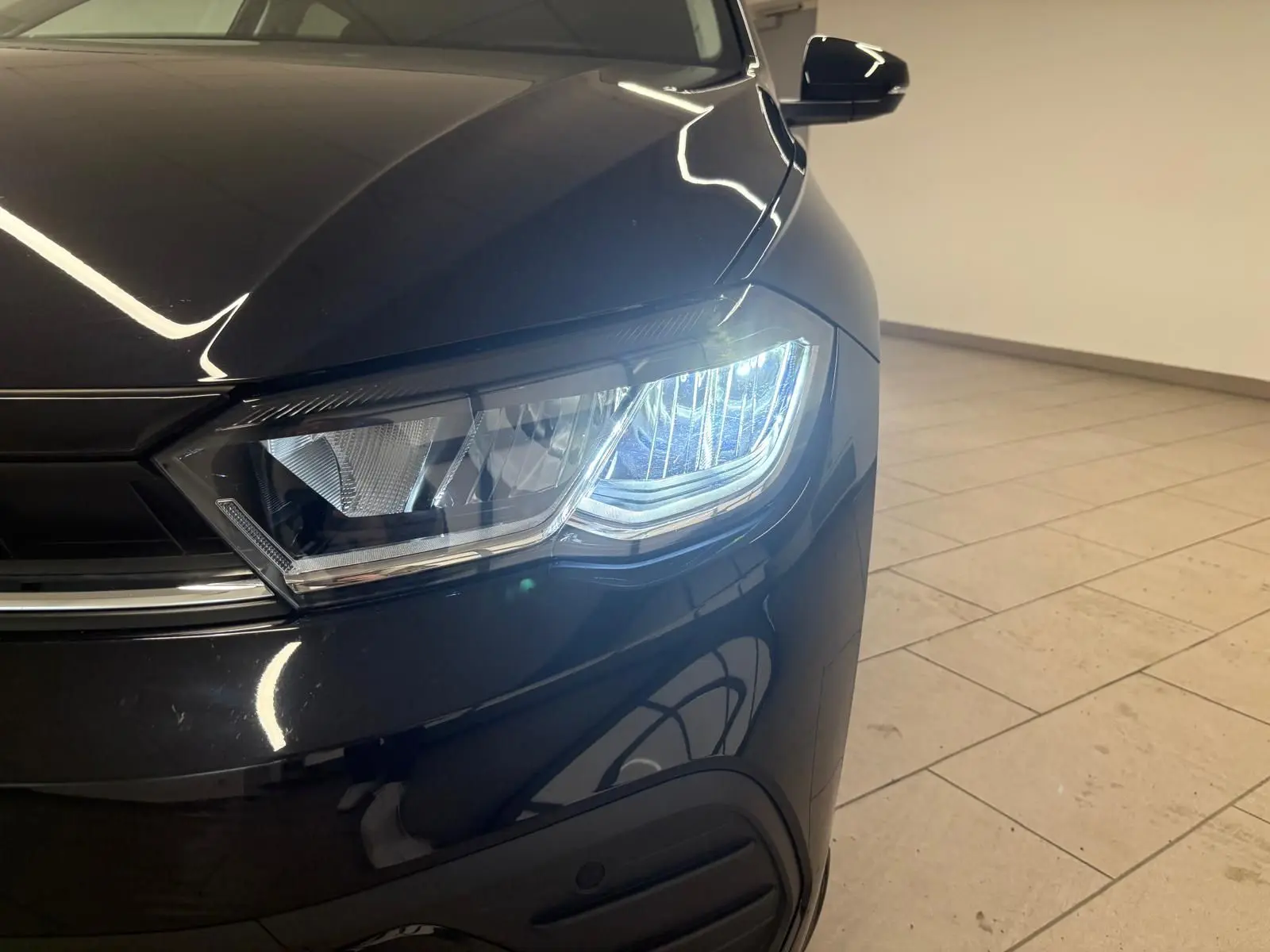 Gros plan sur le phare avant droit allumé d'une Volkswagen Polo noire intense, avec reflets sur la carrosserie.