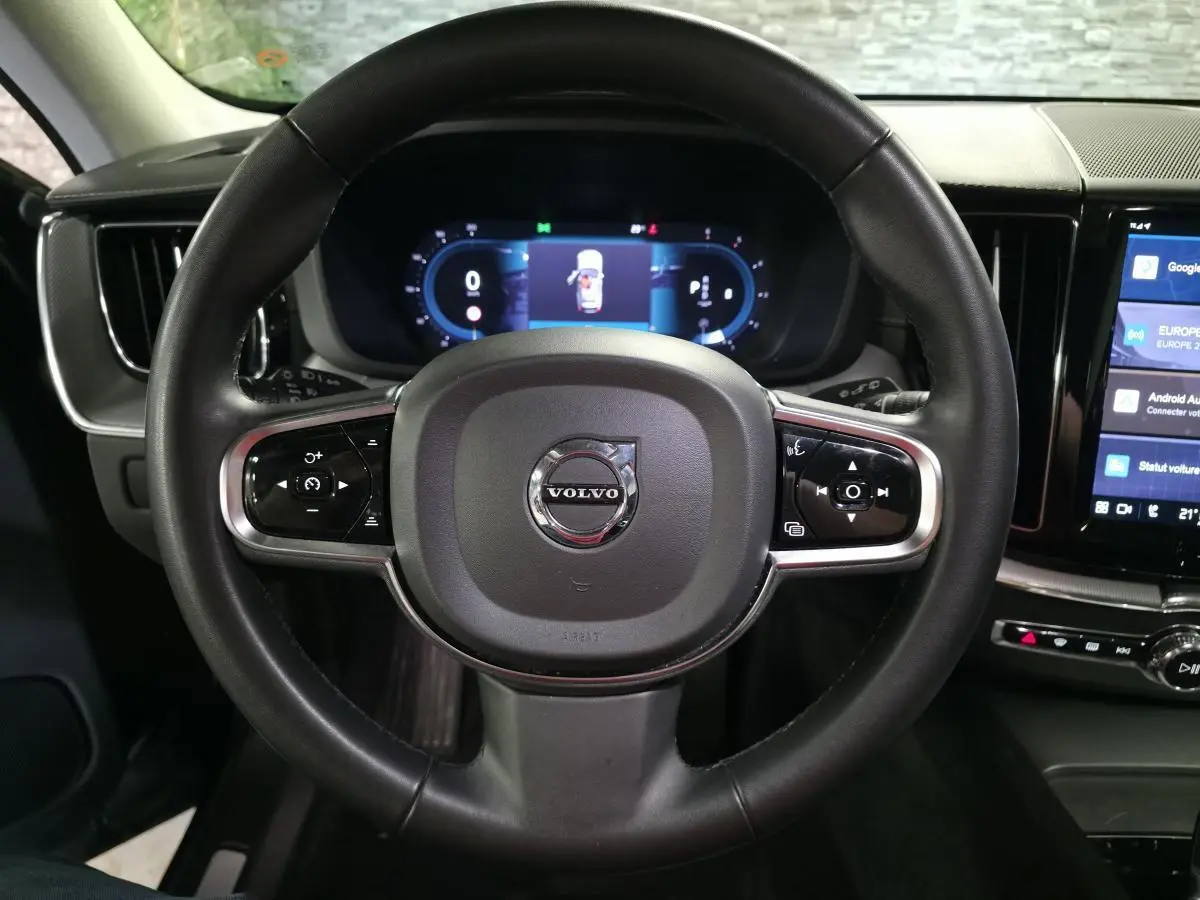 Vue rapprochée du volant noir de la Volvo XC60 2023 avec commandes intégrées et tableau de bord digital en arrière-plan.