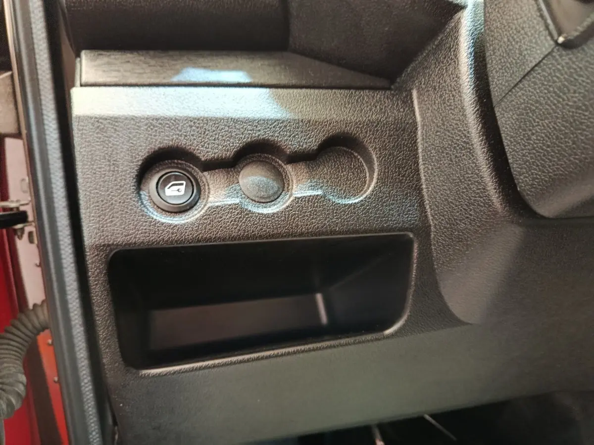 Détail intérieur montrant le bouton d'ouverture du coffre sur la partie gauche du tableau de bord noir de l'AIXAM Ambition rouge.