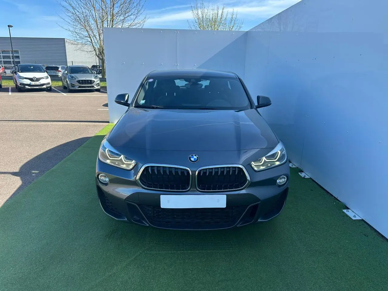 Vue frontale d'un BMW X2 gris foncé avec calandre double haricot et phares LED allumés sur fond urbain.