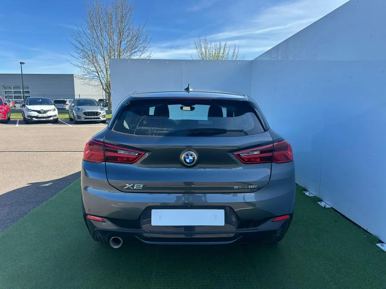 Vue arrière d'une BMW X2 gris foncé avec sortie d'échappement chromée et feux arrière LED, stationnée sur sol vert.