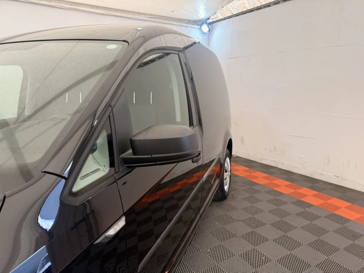 Vue de profil côté gauche d’un Volkswagen Caddy noir 2019 en intérieur avec sol à damier noir et orange.