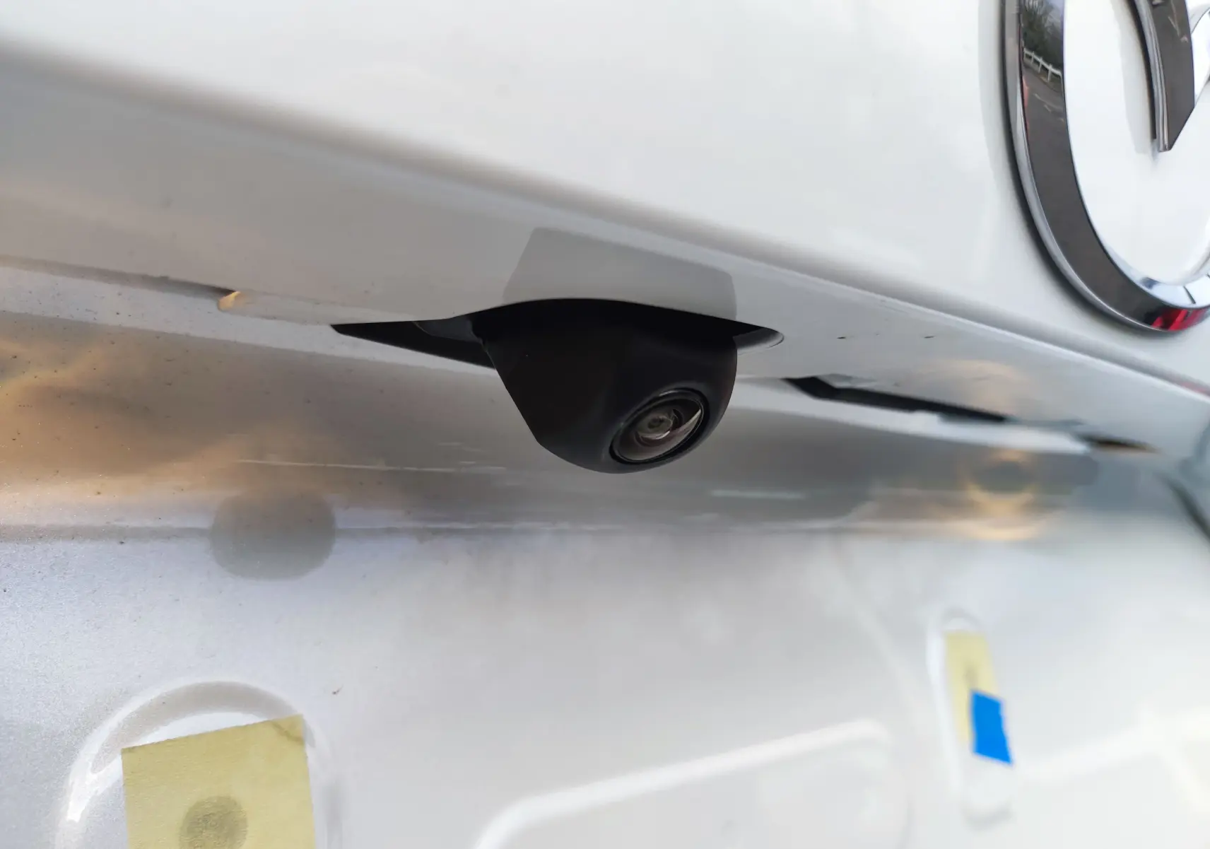 Gros plan sur la caméra de recul noire sous le logo Mazda sur la malle arrière blanche métallisée d'une Mazda2 Hybrid 2024.