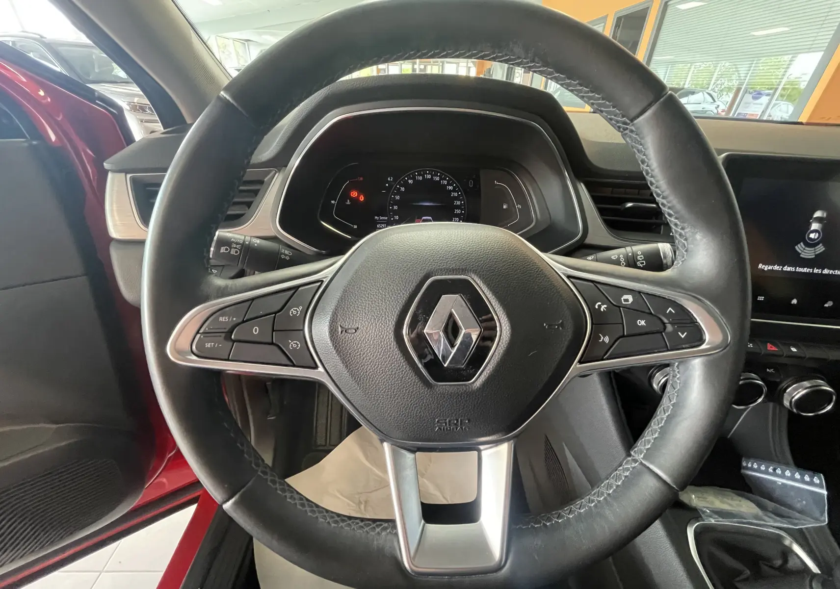 Vue intérieure centrée sur le volant noir du Renault Captur rouge, avec tableau de bord numérique et levier de vitesses manuel visible.