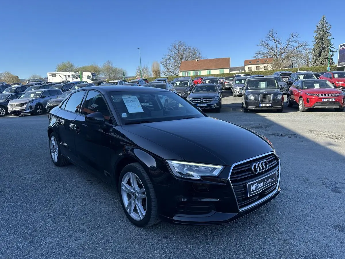 Audi A3 berline noire vue en 3/4 avant droit sur un parking avec plusieurs autres véhicules en arrière-plan.