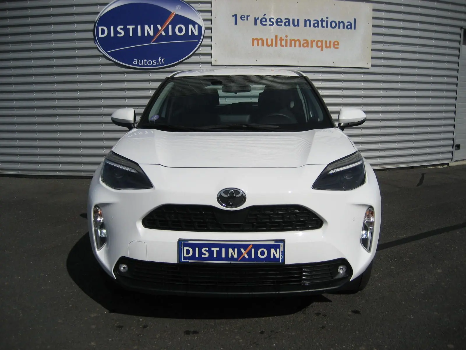 Vue avant d'une Toyota Yaris Cross blanche 2025 avec calandre noire et phares LED sombres, stationnée devant un mur métallique.
