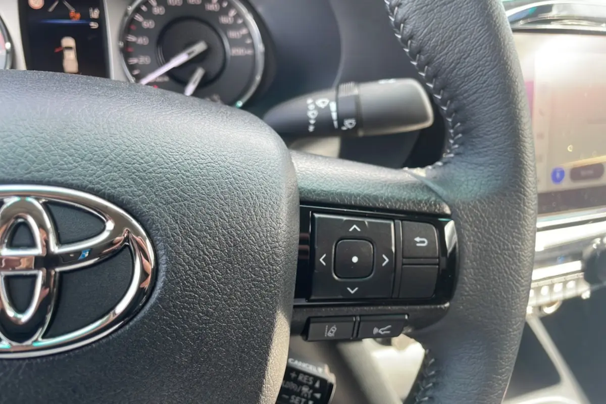 Gros plan sur le volant en cuir noir du Toyota Hilux 2025, avec commandes multifonctions et logo chromé visible.