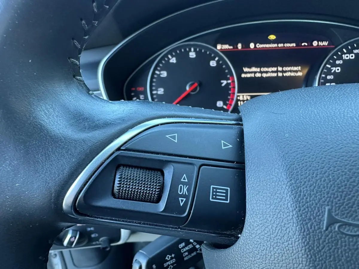 Gros plan sur les commandes gauche du volant cuir d’une Audi A6 2.0 TFSI 2015 avec tableau de bord numérique.