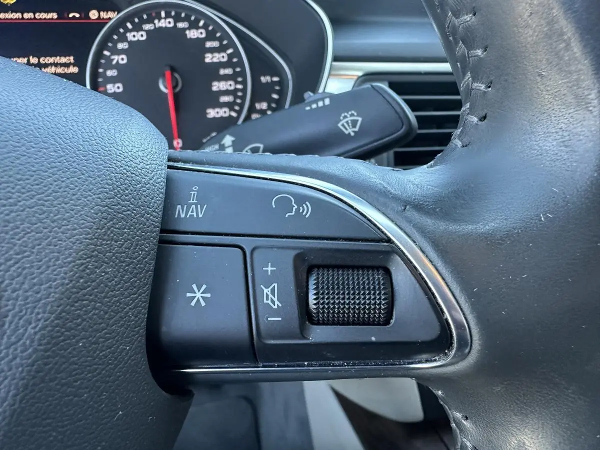 Gros plan sur les commandes vocales et volume au volant cuir noir de l'Audi A6 gris 2015, tableau de bord en arrière-plan.