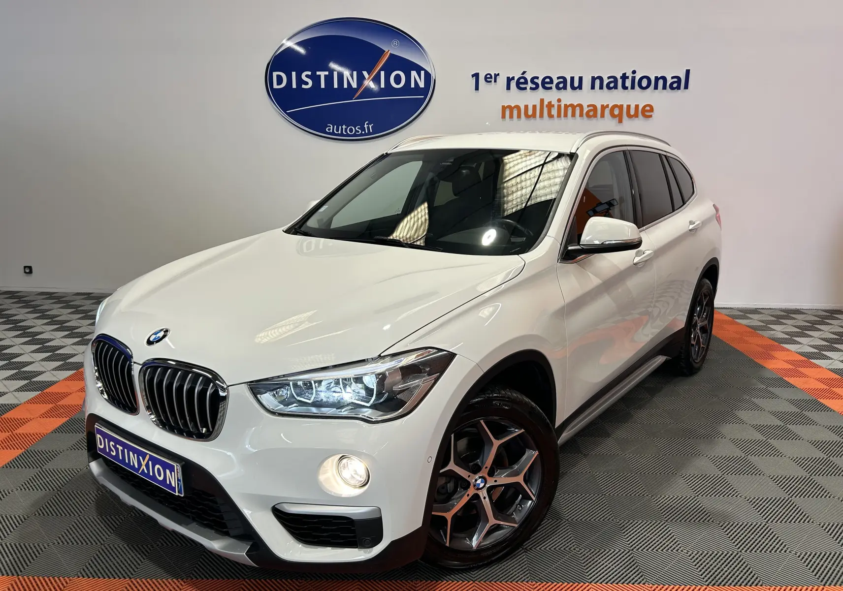 BMW X1 sDrive18iA blanc en 3/4 avant droit avec jantes bi-ton et hayon électrique visible en arrière-plan.