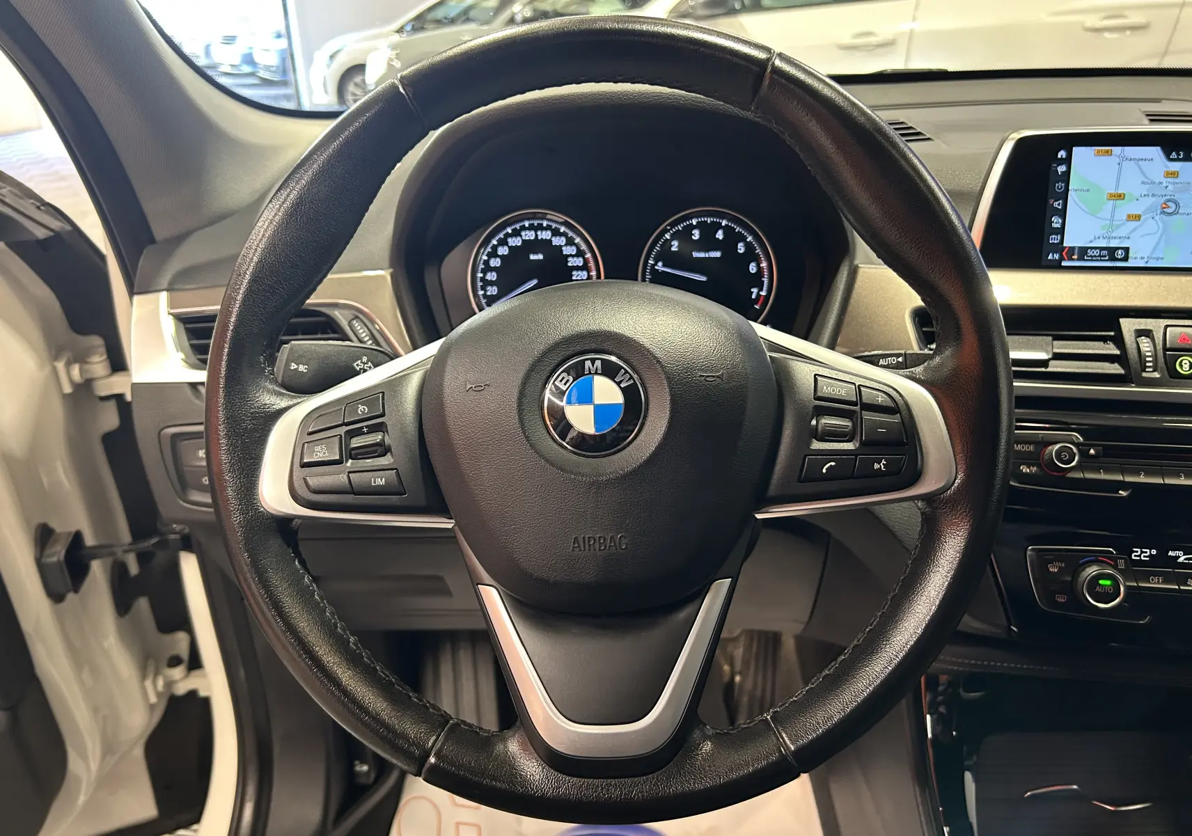 Vue intérieure centrée sur le volant cuir noir d'une BMW X1 blanche avec tableau de bord et écran GPS visible.