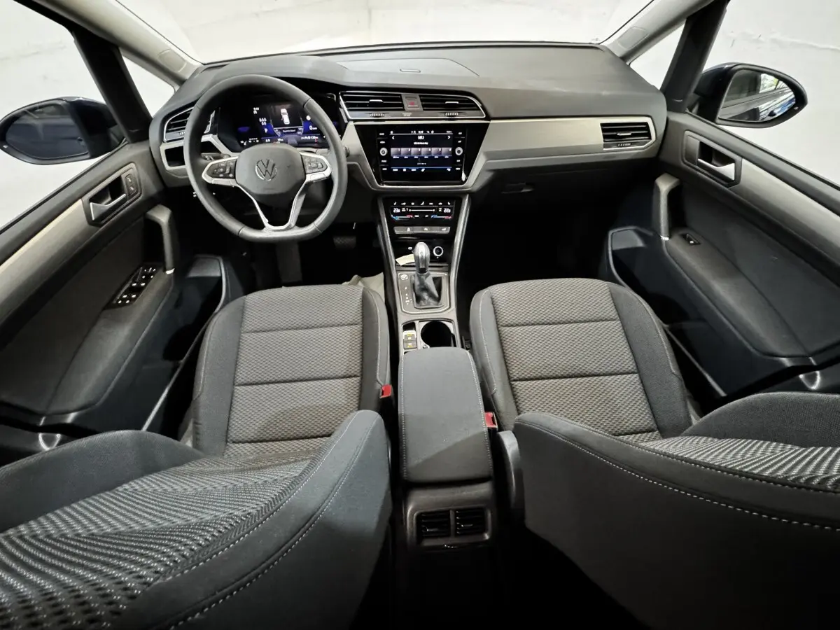 Vue intérieure avant du Volkswagen Touran 2024 avec tableau de bord noir et sièges tissu gris foncé.